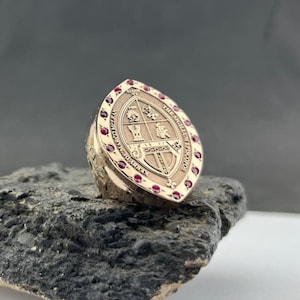 Könnte beinhalten: Ein marquiseförmiger Goldring mit einem zentralen Wappen, das vier Quadranten und ein Turmemblem zeigt. Der Rand ist mit kleinen runden roten Edelsteinen besetzt. Die Metalloberfläche ist mit dem Text THE SIGILLUM EPISCOPOS und KERRY DE MARCO MCLEAN graviert.