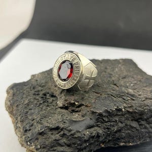 May include: A silver ring with a large, oval, deep red gemstone at the center. The ring's band features the words "THE OHIO STATE UNIVERSITY" around the gemstone and a cross design on the side.