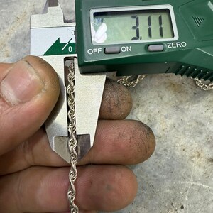 May include: A silver rope chain is measured with a digital caliper. The caliper displays a measurement of 3.11 mm, or approximately 0.12 inches. The chain is held between the caliper jaws, showing its twisted rope design.