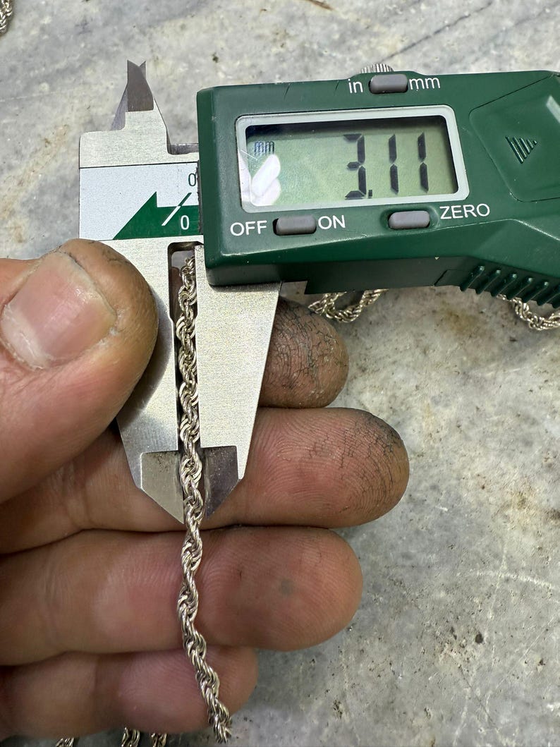May include: A silver rope chain is measured with a digital caliper. The caliper displays a measurement of 3.11 mm, or approximately 0.12 inches. The chain is held between the caliper jaws, showing its twisted rope design.