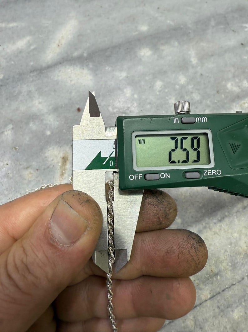 May include: A silver rope chain is measured with a digital caliper. The caliper displays a measurement of 2.59 mm. The chain is held by a hand with visible dirt.