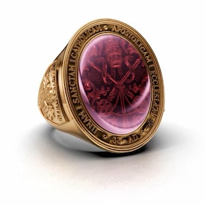May include: A gold ring featuring a large, oval, deep red gemstone set in the center. The metal band is engraved with the Latin text "UNAM SANCTAM CATHOLICAM APOSTOLICAM ECCLESIAM" and "AD 33" around the bezel, with intricate relief carvings on the sides.