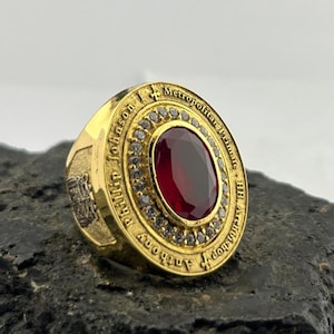 May include: A gold-colored ring with a large, oval, deep red gemstone at the center. The ring is inscribed with text around the gemstone and on the band. Small, clear stones surround the central gem. The ring is set on a dark, textured surface.
