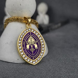 May include: A gold-toned pendant featuring an oval-shaped purple center with the text THE SEAL OF THE BISHOP INTER-FAITH FELLOWSHIP ASSEMBLIES, a mitre, a dove, and a cross. The pendant is bordered by clear stones and hangs from a gold chain.