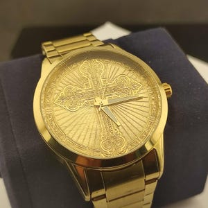 Op de afbeelding: Een goudkleurig analoog polshorloge met een metallic gouden wijzerplaat met een reli&euml;f van een sierlijk kruisontwerp en stralende lijnen. Het horloge heeft een bijpassende goudkleurige metalen schakelarmband en eenvoudige, slanke wijzers.
