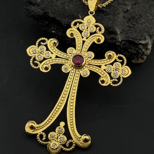 Peut inclure: Pendentif en forme de croix dorée, orné d'un rubis central et de petites pierres claires. La croix, au design élaboré, est suspendue à une chaîne dorée. Le pendentif est présenté sur un fond sombre.
