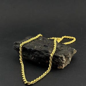 May include: A gold-colored rope chain necklace draped over a dark, textured rock. The chain has a twisted design and is displayed against a black background. The necklace is a jewelry item.