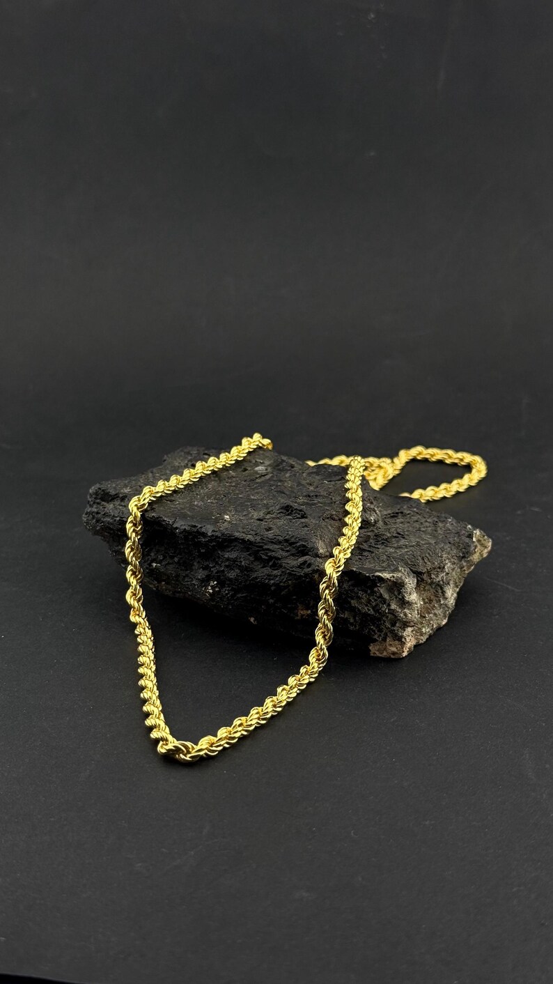 May include: A gold-colored rope chain necklace draped over a dark, textured rock. The chain has a twisted design and is displayed against a black background. The necklace is a jewelry item.