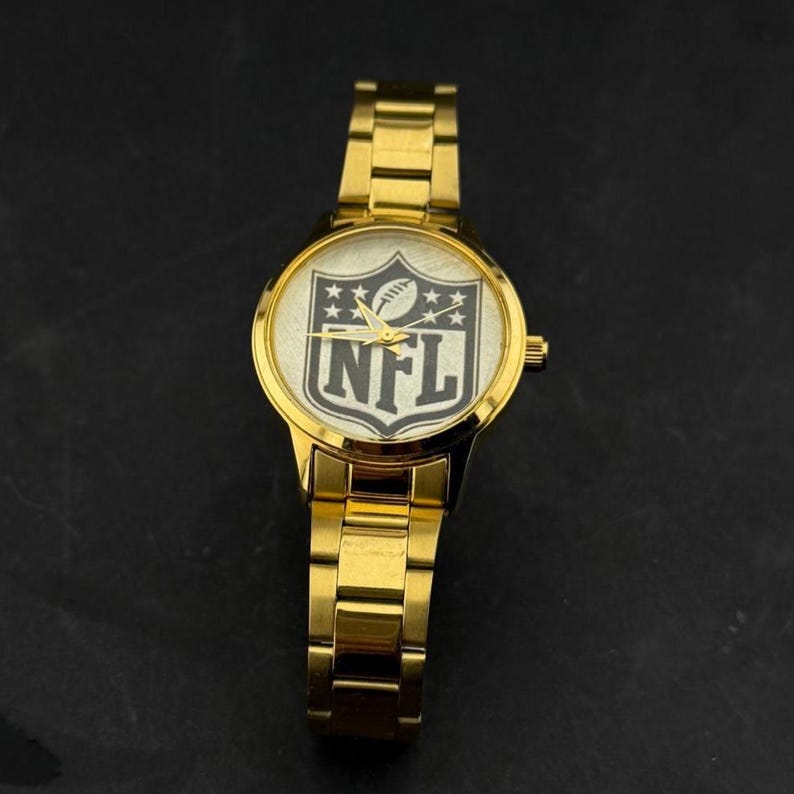 Puede incluir: Un reloj de pulsera dorado con una esfera redonda que muestra el logotipo de la NFL en blanco y negro. El reloj tiene una correa dorada y un bisel dorado. Las manecillas del reloj son doradas. El reloj est&aacute; sobre una superficie oscura.