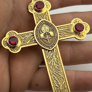 May include: A gold-toned metal cross pendant featuring four round, deep red gemstones set at the ends of each arm. The center displays a circular emblem with a mitre, a sword, and the text THE SEAL OF THE PRESIDING BISHOP INTER FAITH FELLOWSHIP ASSEMBLIES.