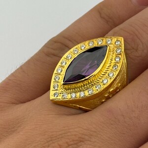 May include: A gold-colored ring featuring a large, oval-shaped, dark purple gemstone. The ring is surrounded by a row of small, clear stones and has a textured band. The ring is a statement piece of jewelry.