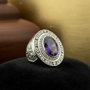 May include: A silver ring featuring a large oval purple gemstone, encircled by smaller clear stones. The ring is inscribed with "THE SEAL OF THE BISHOP JASON NEWTON WRIGHT". The ring rests on a black surface.