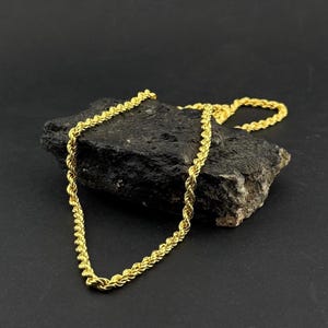 May include: A gold-colored rope chain necklace draped over a dark, textured rock. The chain has a twisted design, reflecting light. The background is a solid black, highlighting the jewelry's details. The necklace is a classic jewelry piece.