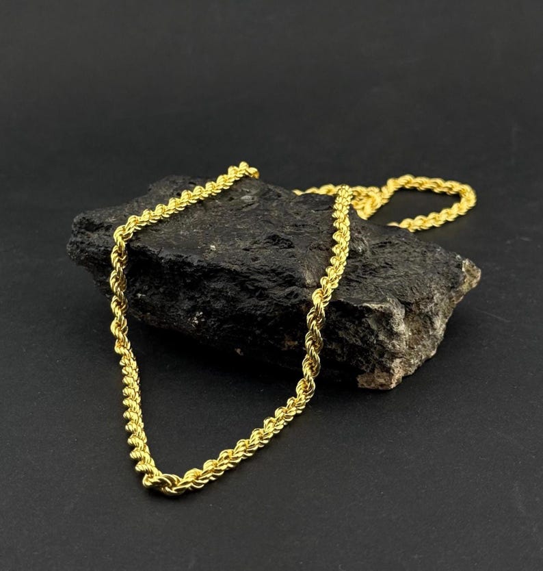 May include: A gold-colored rope chain necklace draped over a dark, textured rock. The chain has a twisted design, reflecting light. The background is a solid black, highlighting the jewelry's details. The necklace is a classic jewelry piece.