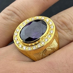 Peut inclure: Bague dorée ornée d'une grande pierre ovale violet foncé. La pierre est entourée de petits brillants. La bague a un anneau épais avec des détails gravés. Un bijou de caractère.