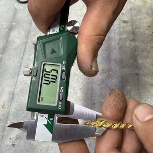 May include: A gold rope chain is measured with a digital caliper. The caliper displays a measurement of 4.03 inches. The chain is held between the caliper jaws, showing its thickness. The caliper is green and silver.