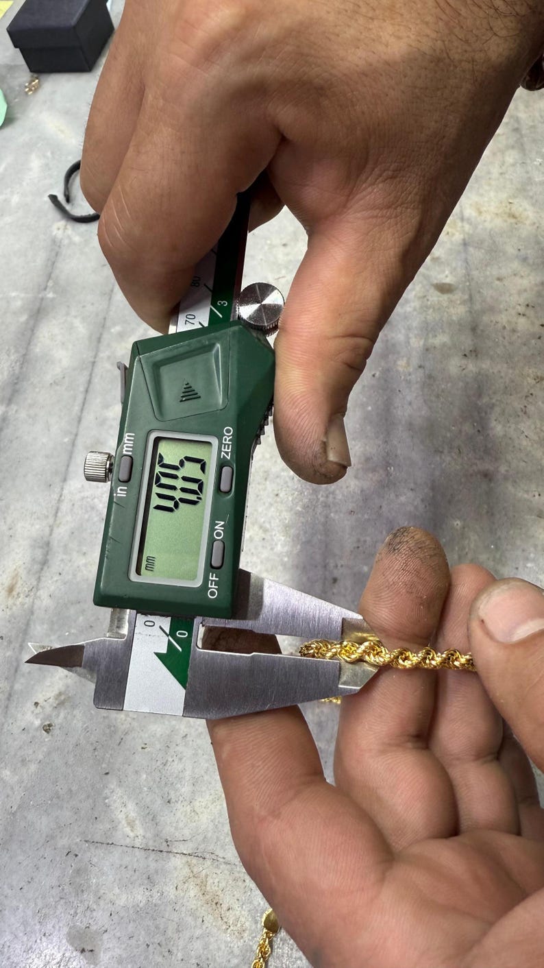 May include: A gold rope chain is measured with a digital caliper. The caliper displays a measurement of 4.03 inches. The chain is held between the caliper jaws, showing its thickness. The caliper is green and silver.