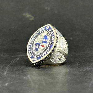May include: A silver-toned ring featuring a shield-shaped face with a blue enamel border inscribed with the text SEAL OF THE OVERSEER SHATANTA SMART. The center displays a four-quadrant crest with symbols including a heart and a book.