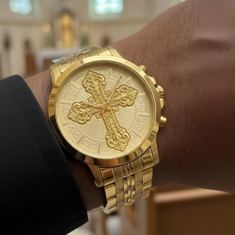 Op de afbeelding: Een goudkleurig analoog horloge met een opvallend, sierlijk, reli&euml;f kruisontwerp op de wijzerplaat. Het horloge heeft een ronde kast, een bijpassende goudkleurige metalen schakelarmband en drie knoppen aan de zijkant, gedragen bij een donkere mouw.