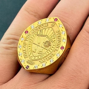 May include: A gold ring featuring a shield-shaped crest with the text SEAL OF THE APOSTLE STEFFON L. TAYLOR MMXXV. The crest includes a compass and other symbols. The border is set with alternating clear white stones and small red stones.