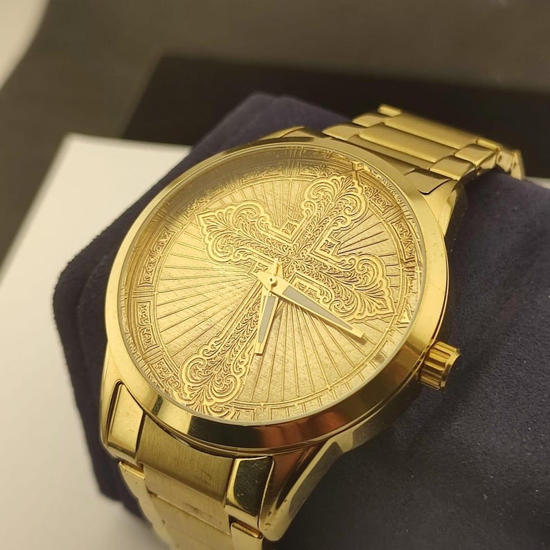 Op de afbeelding: Een goudkleurig analoog polshorloge met een ronde kast en een bijpassende schakelarmband. De wijzerplaat toont een ingewikkeld, in reli&euml;f gemaakt filigraan kruisontwerp gecentreerd tegen een zonnestraalpatroon, met twee dunne, donkere wijzers.