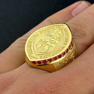 May include: A gold-colored ring with a large oval face featuring an emblem and text. The ring is adorned with small, red gemstones around the face. The text on the ring reads "THE SIGILLUM EPISCOPUS WILLIAMS".