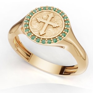 May include: A gold signet ring featuring a circular face with an engraved cross surrounded by intricate scrollwork patterns. The center design is bordered by a halo of small, round, green gemstones set into the polished gold band.