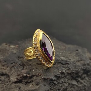 May include: A gold ring featuring a large marquise-cut purple gemstone at the centre, surrounded by a halo of small clear stones. The gold band displays a textured surface with a distinct horseshoe-shaped engraving on the side of the shank.