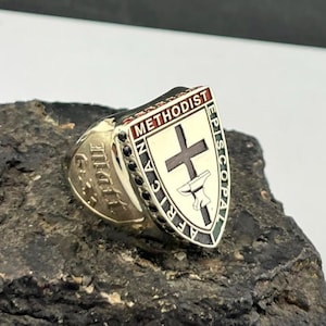 May include: A gold-toned ring featuring a shield-shaped face with the text AFRICAN METHODIST EPISCOPAL surrounding a central cross and anvil emblem. The ring is accented with small black stones along the side and engraved lettering on the shank.