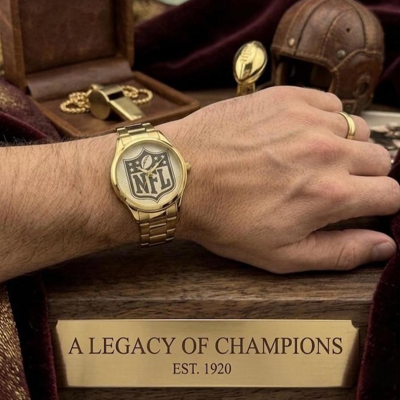 Puede incluir: Reloj de pulsera dorado con el logotipo de la NFL en la esfera. El reloj tiene una correa dorada y se muestra sobre una superficie de madera. El texto "A LEGACY OF CHAMPIONS EST. 1920" est&aacute; grabado en una placa de lat&oacute;n.