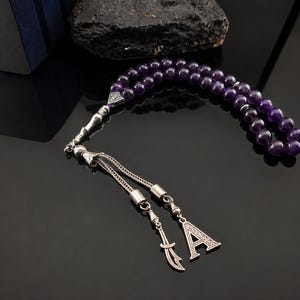 May include: A string of deep purple amethyst prayer beads featuring a silver-toned metal tassel. The tassel includes two dangling charms: one shaped like a curved sword and the other shaped like the capital letter A with an intricate engraved pattern.