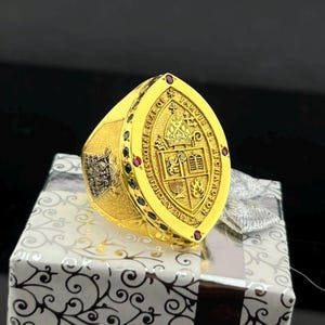 May include: A gold ring featuring an oval face with an embossed crest, a mitre, and the text "THE ARCHIEPISCOPAL SEAL OF JARVIS C. RICHARDSON". The border is accented with small red and green stones, and the side shank displays a detailed engraved emblem.