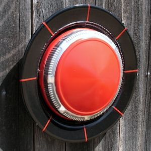 May include: A red and black hubcap with a chrome center ring. The hubcap is mounted on a wooden wall.