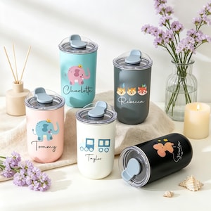 May include: Five cylindrical tumblers with clear lids and blue flip-top closures. Colors include light blue, dark gray, light pink, white, and black. Each features a unique graphic and a name: Charlotte, Rebecca, Tommy, Taylor, and Julian.