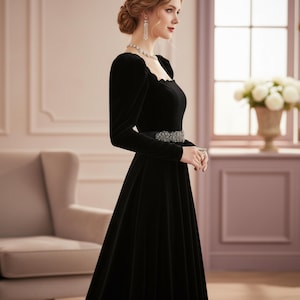May include: A full-length black velvet gown with a square neckline and long sleeves. The dress is cinched at the waist with a sparkling belt. The woman is wearing a necklace and earrings. The background includes a chair and a window, with a vase of flowers.