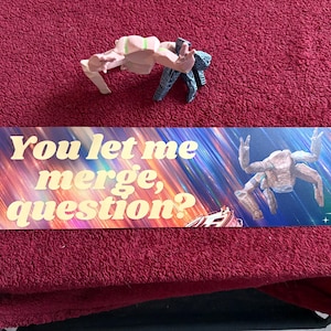 Puede incluir: Un letrero rectangular con el texto "You let me merge, question?" en letras doradas. El fondo presenta un diseño abstracto colorido con una figura de cangrejo. Dos pequeñas figuras de cangrejo están encima.