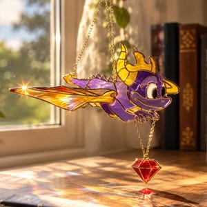 Puede incluir: Un atrapasueños de vidrieras que representa un dragón morado con cuernos amarillos y alas naranjas, suspendido por cadenas doradas. El dragón sostiene una gema roja facetada en forma de diamante que cuelga de una delicada cadena dorada.