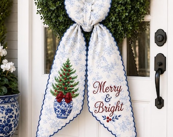 May include: A green boxwood wreath adorned with a large fabric bow featuring a blue and white toile pattern. The bow has two long tails, one displaying a potted Christmas tree graphic and the other the red text Merry & Bright with scalloped edges.