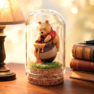 May include: A Winnie the Pooh figurine inside a glass dome. The bear is reaching into a honey pot, with honey dripping down. The figurine sits on a bed of green grass and decorative rocks. A lamp and books are in the background.