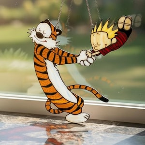 Könnte beinhalten: Ein Buntglas-Suncatcher, der einen Cartoon-Tiger mit orangefarbenen und schwarzen Streifen zeigt, der einen kleinen Jungen an den Händen hält. Das Stück ist an dünnen Metallketten aufgehängt und weist durchscheinende Farben auf.