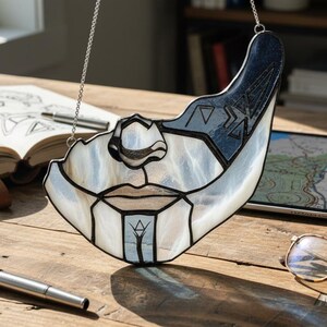 May include: A stained glass suncatcher in the shape of a stylized face, with a silver chain for hanging. The design features white, blue, and black glass pieces, with geometric patterns. The suncatcher is displayed on a wooden surface.