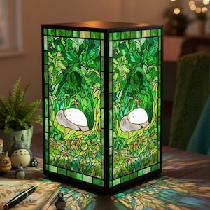 May include: A stained glass lamp with a black frame. The lamp features a green forest scene with white and gray characters. The lamp is on a wooden table with a notebook, pen, and figurines.