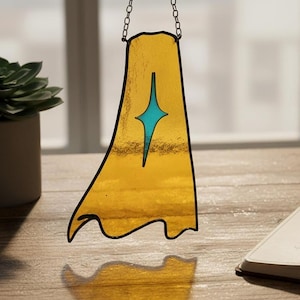 以下が含まれることがあります： 金色のステンドグラスのサンキャッチャー。ケープの形をしており、ターコイズ色の星のデザインが施されています。チェーンで吊り下げられており、窓の前に飾られています。多肉植物とノートが背景にあります。