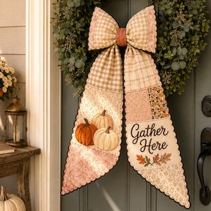 May include: A circular green foliage wreath featuring a large fabric bow with long, scalloped-edge tails. The bow displays a beige gingham pattern, pumpkin illustrations, and the text "Gather Here" with small autumn leaf accents.