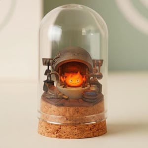 Puede incluir: Una c&uacute;pula de cristal decorativa con una escena en miniatura de un esp&iacute;ritu de fuego naranja brillante. La base es de corcho, y el interior detallado incluye un horno y herramientas. La c&uacute;pula transparente ofrece una vista completa de la escena.