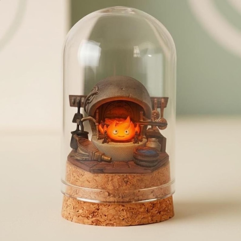 Puede incluir: Una c&uacute;pula de cristal decorativa con una escena en miniatura de un esp&iacute;ritu de fuego naranja brillante. La base es de corcho, y el interior detallado incluye un horno y herramientas. La c&uacute;pula transparente ofrece una vista completa de la escena.
