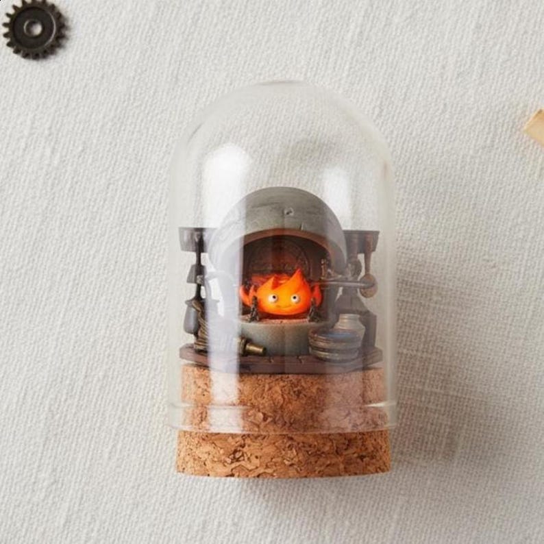 Puede incluir: Un diorama decorativo en miniatura con un personaje ardiente dentro de una c&uacute;pula de cristal. El personaje naranja con una cara sonriente est&aacute; dentro de un horno en miniatura detallado. La base es de corcho y la c&uacute;pula es transparente.