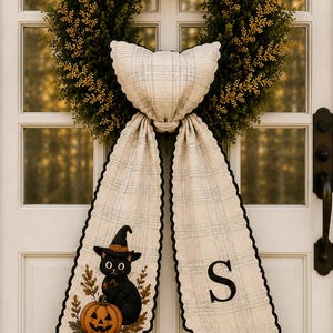 May include: A green wreath with gold accents features a hanging fabric decoration in a cream and grey plaid pattern. The fabric has two long tails, one displaying a black cat wearing a witch hat next to a pumpkin, and the other featuring the letter S.