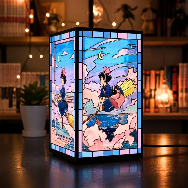 May include: A rectangular stained-glass lamp featuring a young girl on a broomstick, set against a sky with clouds and a crescent moon. The lamp has a black frame with pink, blue, and purple accents. The artwork is reminiscent of a popular animated film.
