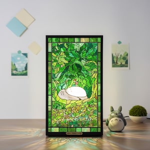 May include: A stained glass lamp featuring a green forest scene with a white creature resting. The lamp has a black frame and the text "Retro Tatten Tang" at the bottom. A small Totoro figure and a small plant are next to the lamp.